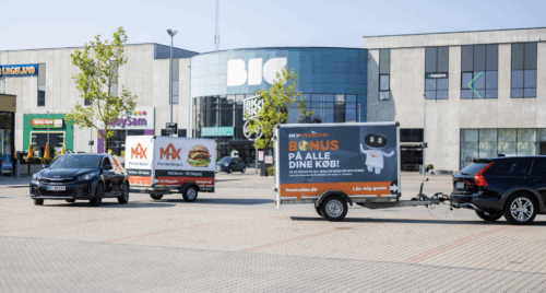 Two cars pulling branded Freetrailer trailers in front of the BIG Shopping center in Herlev, Denmarkâ€”one promoting MAX Burgers and the other POWER. A visual example of how businesses can increase exposure and foot traffic through branded trailers.