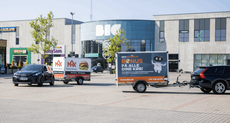 Two cars pulling branded Freetrailer trailers in front of the BIG Shopping center in Herlev, Denmarkâ€”one promoting MAX Burgers and the other POWER. A visual example of how businesses can increase exposure and foot traffic through branded trailers.